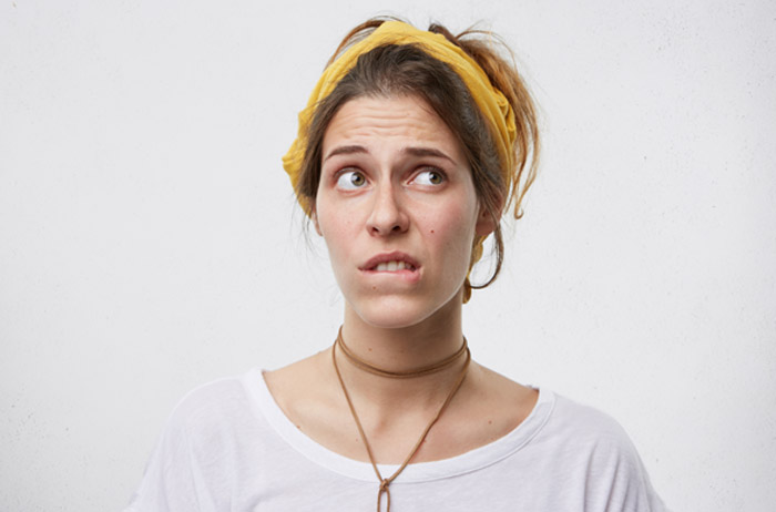 Junge Frau mit braunen Haaren, gelbem Stirnband und grauem T-Shirt vor grauem Hintergrund schaut nachdenklich fragend und verängstigt zur Seite.