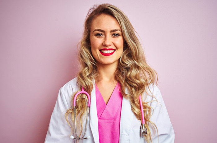 Ärztin vor hell-lila Hintergrund mit langen gelockten blonden Haaren im weißen Kittel und Stethoskop, sie lächelt mit Freude und Selbstbewusstsein den Kooperations-Partnern zu. 