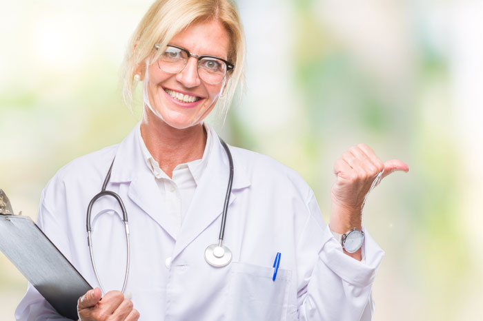 Freundlich lächelnde Ärztin mit blonden Haare und Brille, im Kittel mit Stethoskop und Schreibbrett vor gelb-grünen Hintergrund, hebt linke Hand in Schulterhöhe und streckt Daumen seitlich hoch.