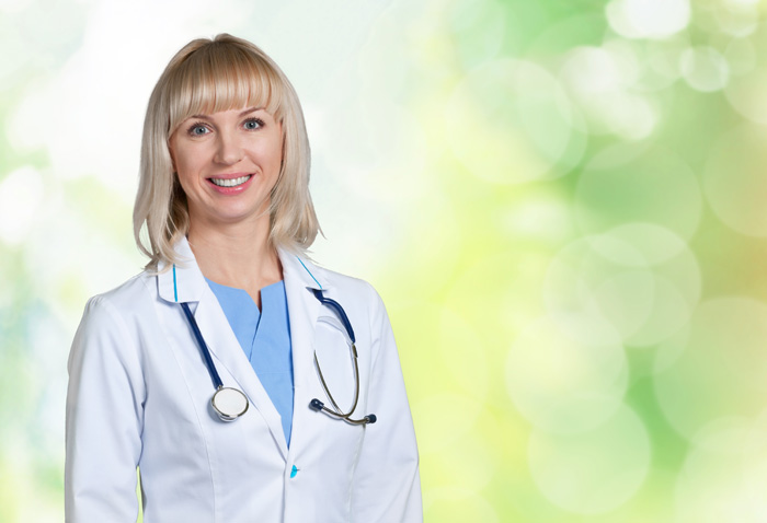 Junge blonde Ärztin mit Kittel und Stethoskop mit grün-gelbem Hintergrund schaut optimistisch und freundlich selbstbewusst in die Kamera.