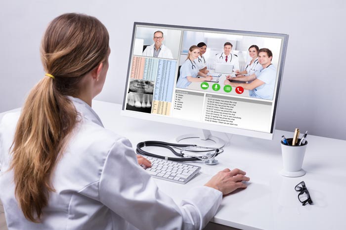 Ärztin mit langen blonden Haaren als  Pferdeschwanz im Kittel sitz vor weißem Schreibtisch mit Bildschirm, auf dem Ärzte als Team-Konferenz  zu sehen sind.