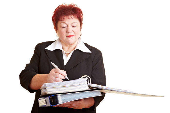 Ältere Frau mit kurzen roten Haaren vor einem weißen Hintergrund macht sich prüfend Aktennotizen.