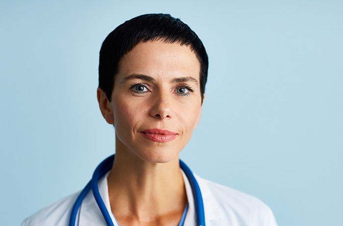 Ärztin mit sehr kurzen schwarzen Haaren in weißem Kittel vor blauem Hintergrund schaut überzeugend aus dem Bild auf den Betrachter.