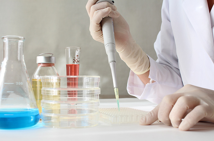 Arm und Hand mit weißem Gummihandschuh und Labor-Pipette, Glasflaschen mit blauer, gelber und roter Flüssigkeit, Laborschalen aus Glas